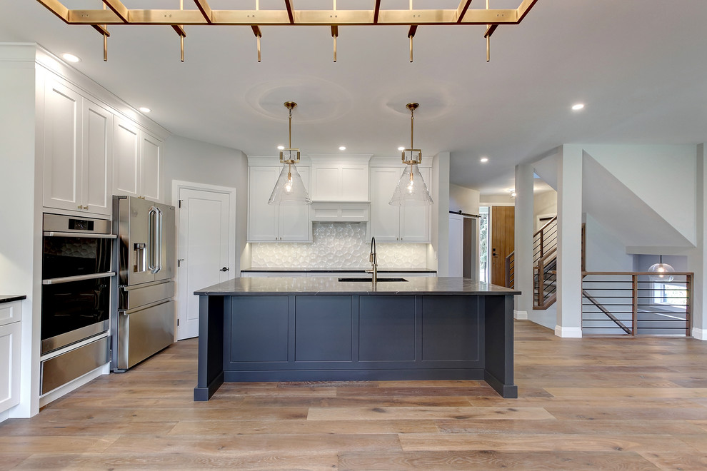 Open concept kitchen - mid-sized transitional l-shaped medium tone wood floor and brown floor open concept kitchen idea in Edmonton with an undermount sink, recessed-panel cabinets, white cabinets, quartz countertops, white backsplash, ceramic backsplash, stainless steel appliances, an island and black countertops
