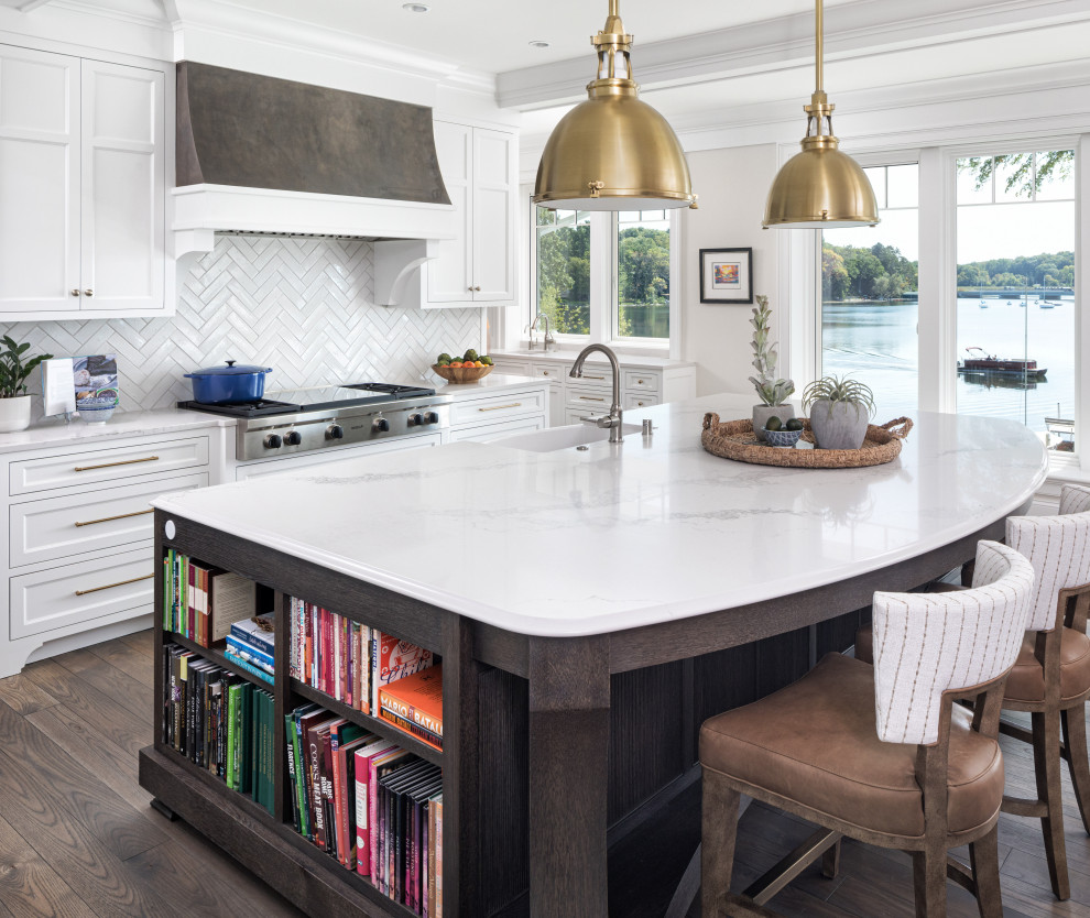 This is an example of a beach style kitchen in Minneapolis with a farmhouse sink, shaker cabinets, white cabinets, white splashback, stainless steel appliances, dark hardwood floors, with island, brown floor and white benchtop.