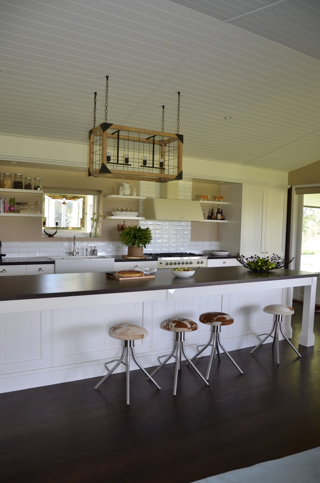 Large country galley open plan kitchen in Melbourne with a farmhouse sink, shaker cabinets, white cabinets, wood benchtops, white splashback, ceramic splashback, white appliances, dark hardwood floors, with island and grey benchtop.