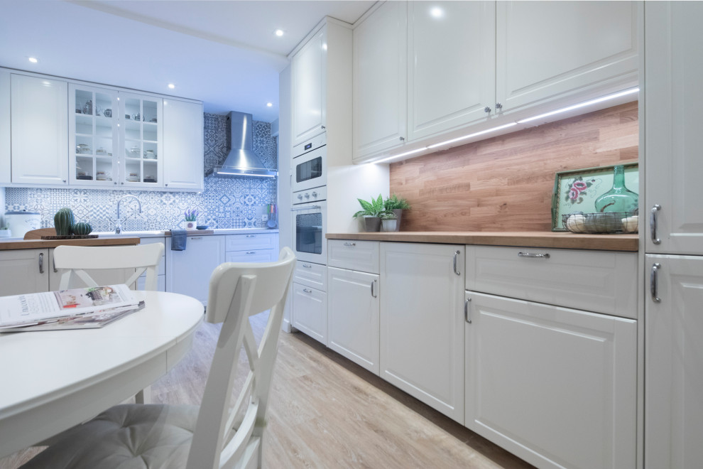Photo of a mid-sized country u-shaped eat-in kitchen in Other with a double-bowl sink, raised-panel cabinets, white cabinets, wood benchtops, blue splashback, ceramic splashback, white appliances, vinyl floors, with island, brown floor, brown benchtop and recessed.
