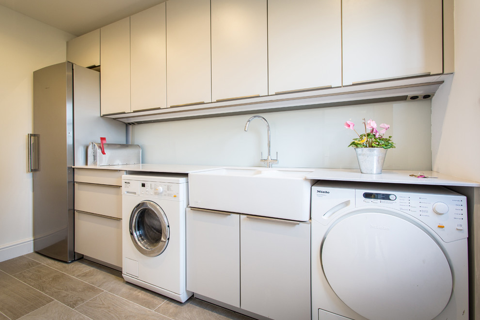 Inspiration for a small contemporary single-wall dedicated laundry room in Hampshire with grey cabinets, metallic splashback, mirror splashback, a farmhouse sink, flat-panel cabinets, solid surface benchtops, white walls and a side-by-side washer and dryer.