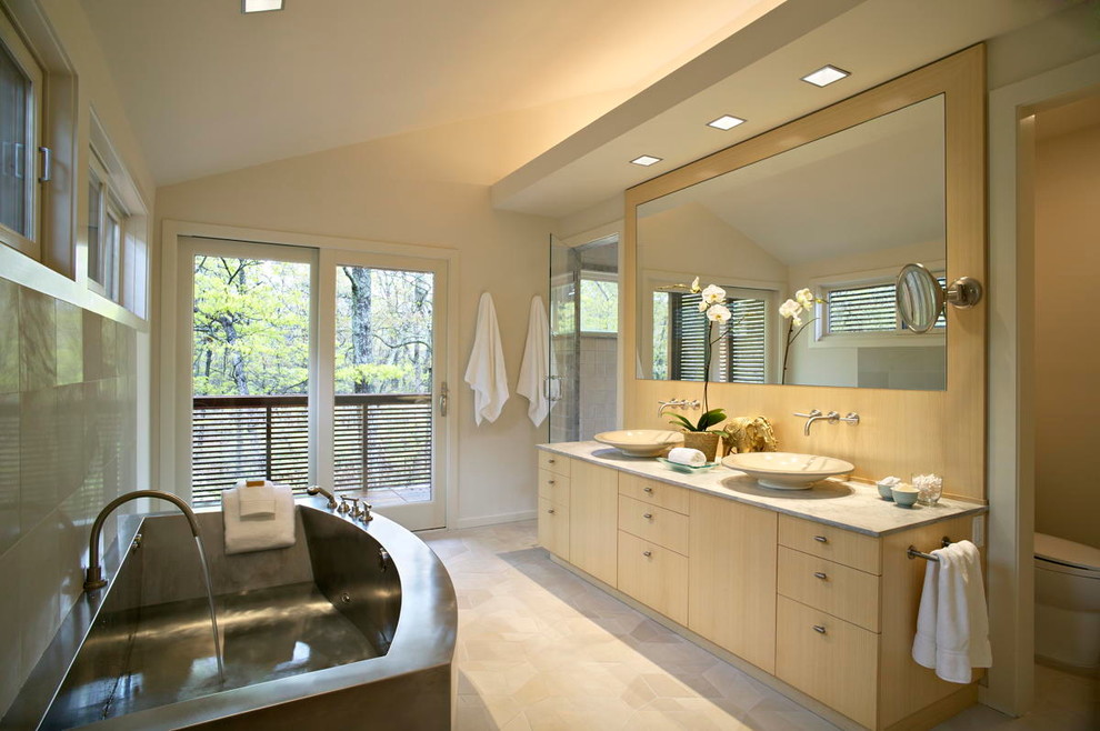 This is an example of a mid-sized modern kids bathroom in New York with flat-panel cabinets, light wood cabinets, a freestanding tub, a corner shower, a one-piece toilet, white tile, stone tile, white walls, limestone floors, a vessel sink, marble benchtops, beige floor and a shower curtain.