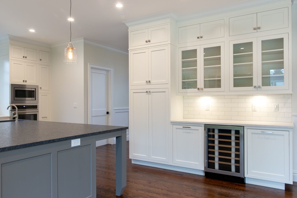 ニューヨークのトランジショナルスタイルのおしゃれなキッチン (エプロンフロントシンク、シェーカースタイル扉のキャビネット、白いキャビネット、御影石カウンター、白いキッチンパネル、磁器タイルのキッチンパネル、シルバーの調理設備、濃色無垢フローリング、白いキッチンカウンター) の写真