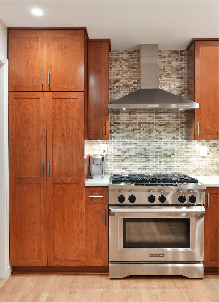 Photo of a mid-sized transitional l-shaped separate kitchen in DC Metro with an undermount sink, glass-front cabinets, medium wood cabinets, marble benchtops, multi-coloured splashback, glass tile splashback, stainless steel appliances, light hardwood floors, no island, beige floor and white benchtop.