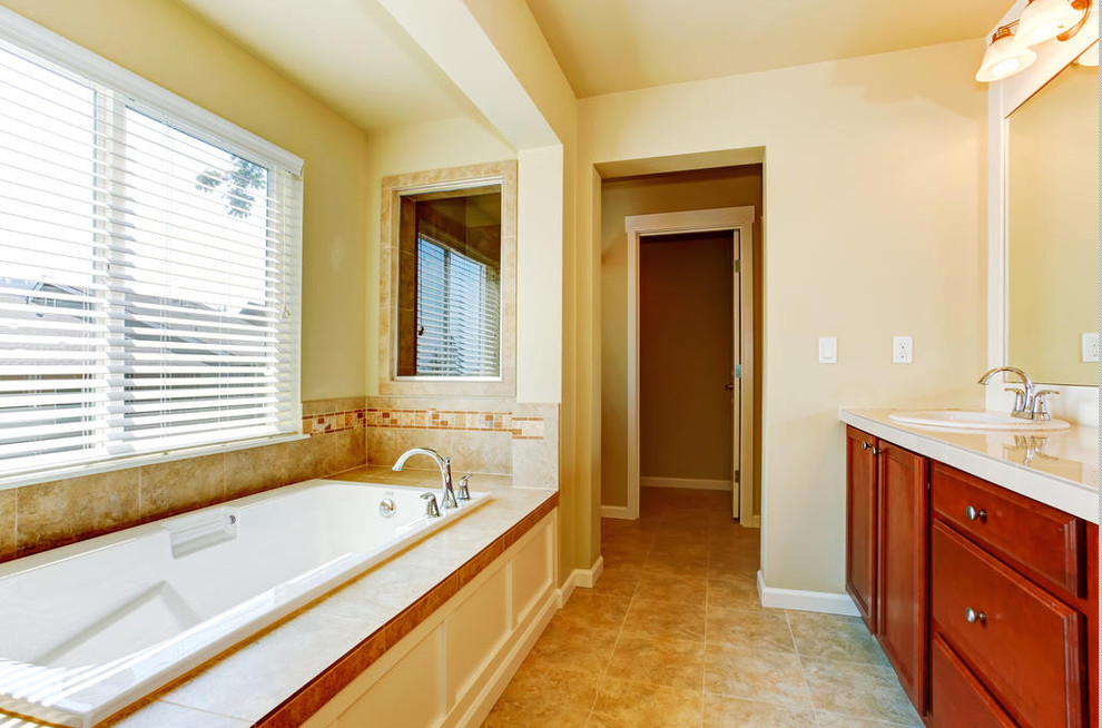 Photo of a mid-sized master bathroom in New York with a drop-in sink, dark wood cabinets, solid surface benchtops, a drop-in tub, beige tile, ceramic tile, beige walls and ceramic floors.