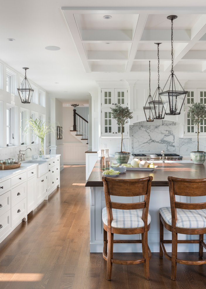 This is an example of a beach style kitchen in Boston with a farmhouse sink, white cabinets, white splashback, stone slab splashback, stainless steel appliances, medium hardwood floors, with island, white benchtop and recessed-panel cabinets.