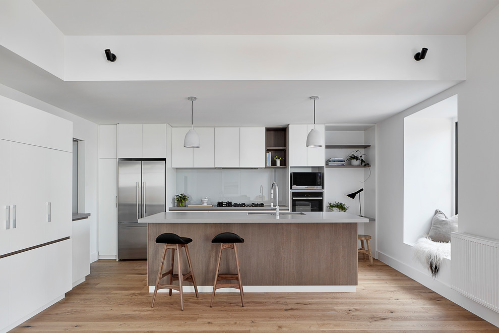 Mid-sized contemporary l-shaped open plan kitchen in Melbourne with an undermount sink, flat-panel cabinets, grey splashback, stainless steel appliances, light hardwood floors, with island, brown floor, quartz benchtops, glass sheet splashback, grey benchtop and vaulted.