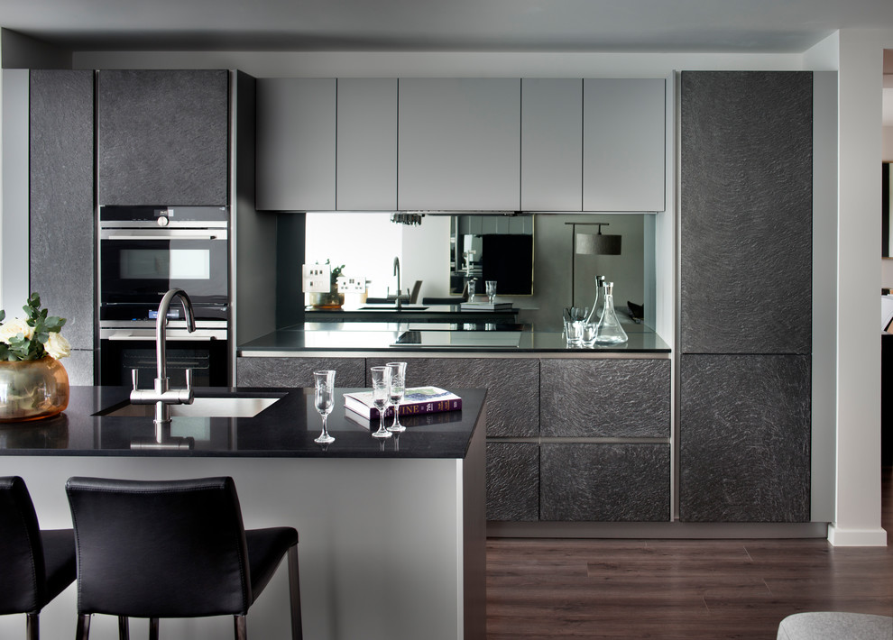 Photo of a mid-sized contemporary galley kitchen in Hertfordshire with an undermount sink, flat-panel cabinets, grey cabinets, mirror splashback, stainless steel appliances, with island and brown floor.