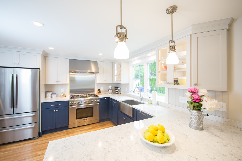 Photo of a beach style eat-in kitchen in Boston with a farmhouse sink, shaker cabinets, blue cabinets, quartz benchtops, grey splashback, timber splashback, stainless steel appliances, medium hardwood floors, a peninsula and white benchtop.