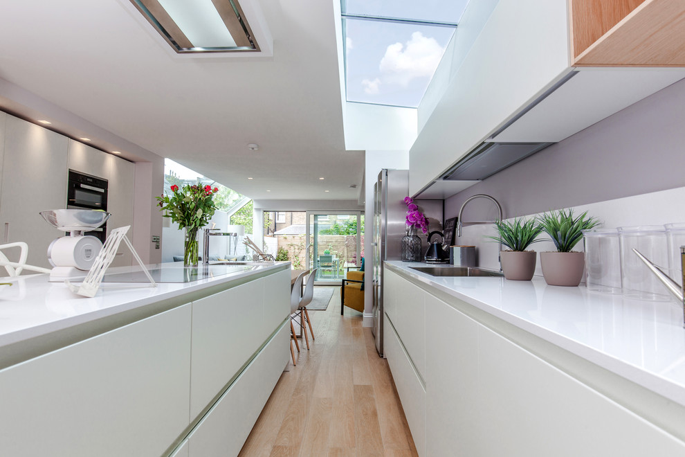 Inspiration for a large contemporary galley open plan kitchen in London with an integrated sink, flat-panel cabinets, white cabinets, quartzite benchtops, white splashback, black appliances, light hardwood floors and with island.
