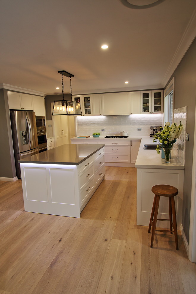 This is an example of a large eclectic l-shaped kitchen pantry in Sydney with an undermount sink, shaker cabinets, white cabinets, quartz benchtops, white splashback, subway tile splashback, stainless steel appliances, light hardwood floors, with island and brown floor.