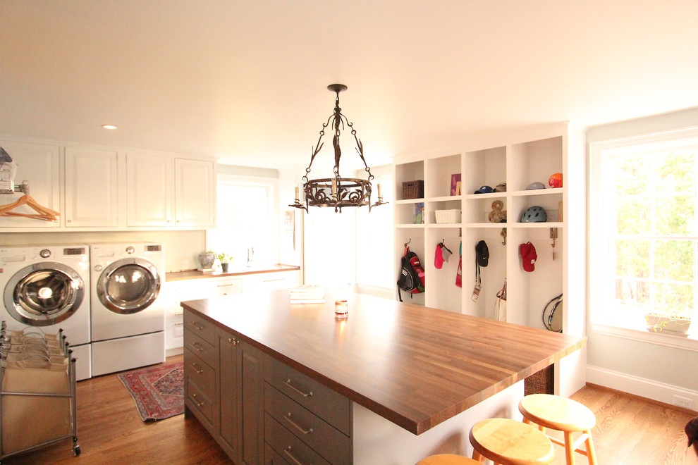 Photo of a large transitional l-shaped utility room in Other with a drop-in sink, raised-panel cabinets, white cabinets, wood benchtops, grey walls, medium hardwood floors, a side-by-side washer and dryer and brown floor.