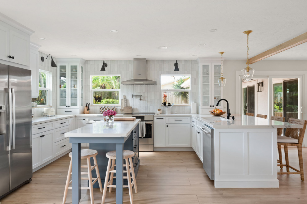 This is an example of a mid-sized beach style u-shaped eat-in kitchen in Tampa with an undermount sink, shaker cabinets, white cabinets, quartz benchtops, white splashback, porcelain splashback, stainless steel appliances, medium hardwood floors, with island, brown floor and white benchtop.