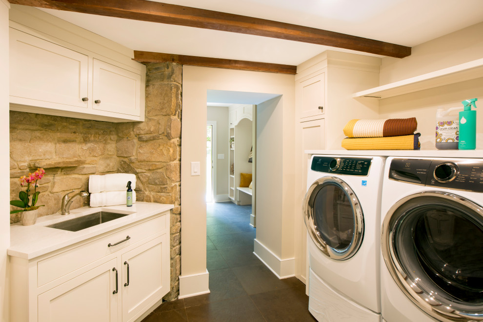 This is an example of a small country u-shaped dedicated laundry room in New York with an undermount sink, recessed-panel cabinets, white cabinets, quartz benchtops, porcelain floors, a side-by-side washer and dryer, grey floor, white benchtop, exposed beam and brick walls.