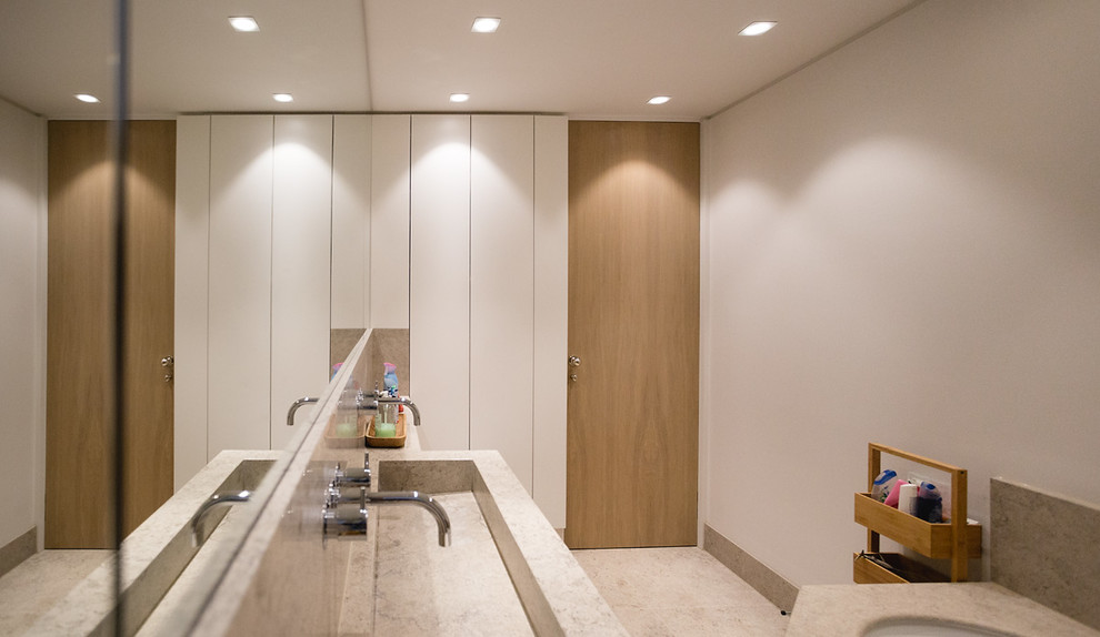 This is an example of a mid-sized contemporary kids bathroom in London with flat-panel cabinets, light wood cabinets, a drop-in tub, a shower/bathtub combo, a wall-mount toilet, beige tile, stone slab, white walls, limestone floors, a trough sink, limestone benchtops, beige floor, a hinged shower door and beige benchtops.