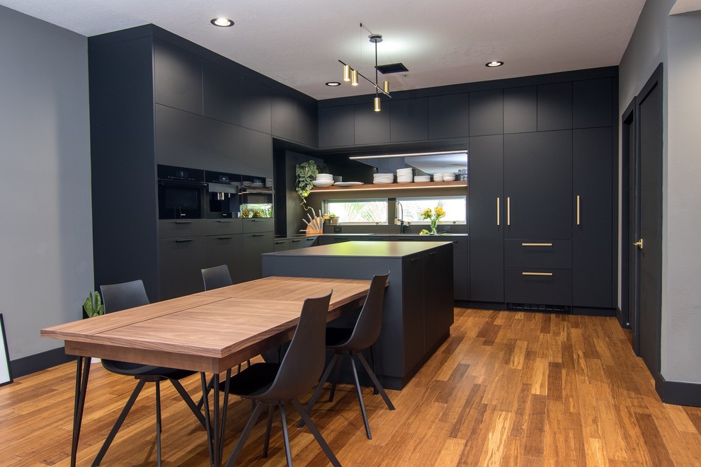 Example of a minimalist l-shaped medium tone wood floor and brown floor eat-in kitchen design with flat-panel cabinets, black cabinets, paneled appliances, an island and black countertops