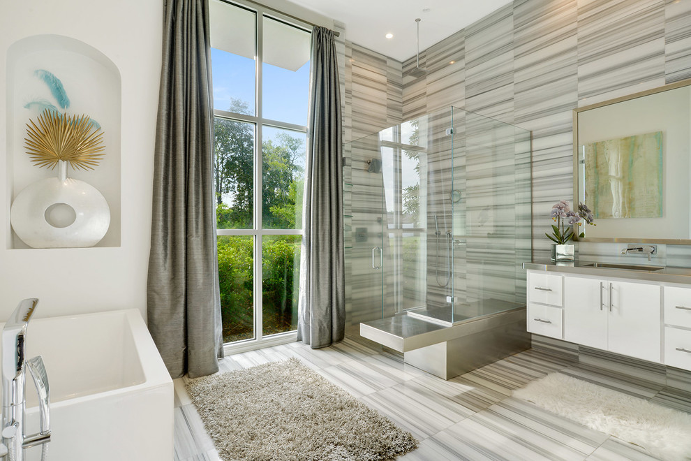 This is an example of a large modern master bathroom in New Orleans with flat-panel cabinets, white cabinets, a freestanding tub, a corner shower, gray tile, stone tile, white walls, marble floors, an integrated sink, stainless steel benchtops, grey floor and a hinged shower door.