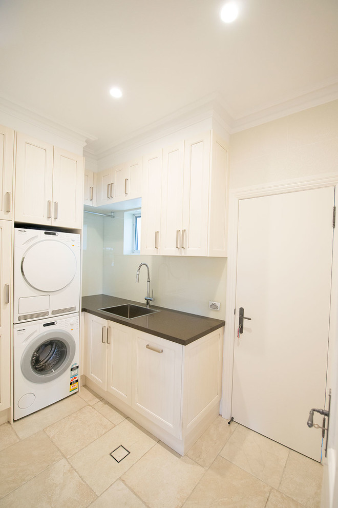 This is an example of a large traditional l-shaped laundry room in Sydney with beige cabinets, white walls, a stacked washer and dryer, shaker cabinets, quartz benchtops, a farmhouse sink, beige splashback, glass sheet splashback and travertine floors.