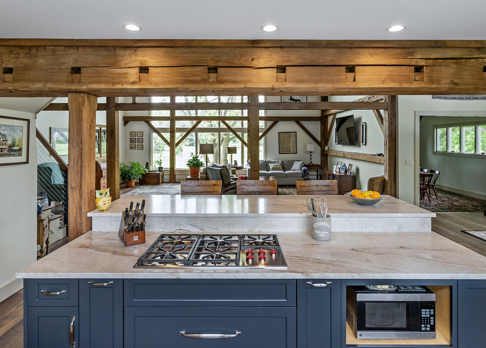 Mid-sized country galley open plan kitchen in Philadelphia with a farmhouse sink, shaker cabinets, blue cabinets, quartz benchtops, white splashback, porcelain splashback, stainless steel appliances, medium hardwood floors, with island, brown floor, white benchtop and exposed beam.