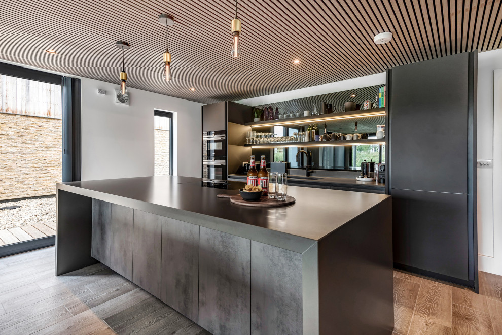 Large contemporary grey and black single-wall open plan kitchen in Gloucestershire with a submerged sink, flat-panel cabinets, grey cabinets, composite countertops, grey splashback, glass sheet splashback, integrated appliances, an island, brown floors, grey worktops, a wood ceiling and feature lighting.