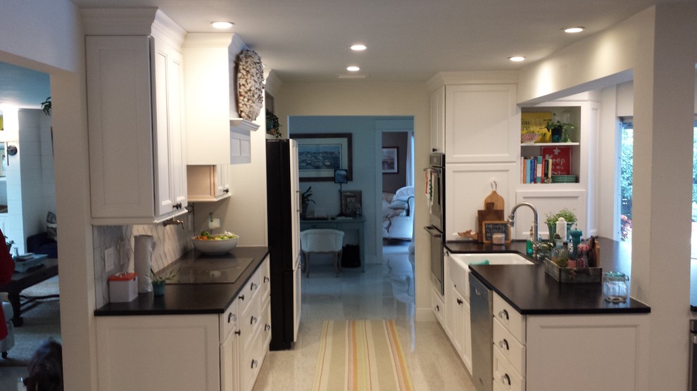 Transitional galley kitchen photo in Tampa with a farmhouse sink, recessed-panel cabinets, white cabinets, granite countertops, beige backsplash, glass tile backsplash, paneled appliances and black countertops