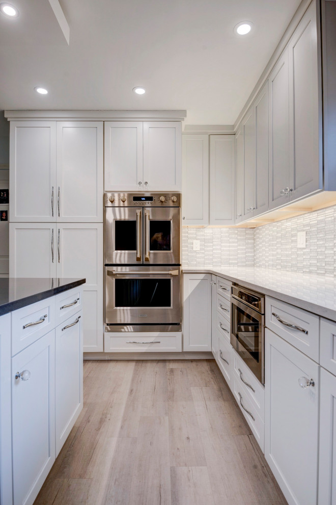 Example of a large transitional u-shaped light wood floor and beige floor open concept kitchen design in New York with an undermount sink, shaker cabinets, white cabinets, quartzite countertops, white backsplash, ceramic backsplash, stainless steel appliances, an island and white countertops