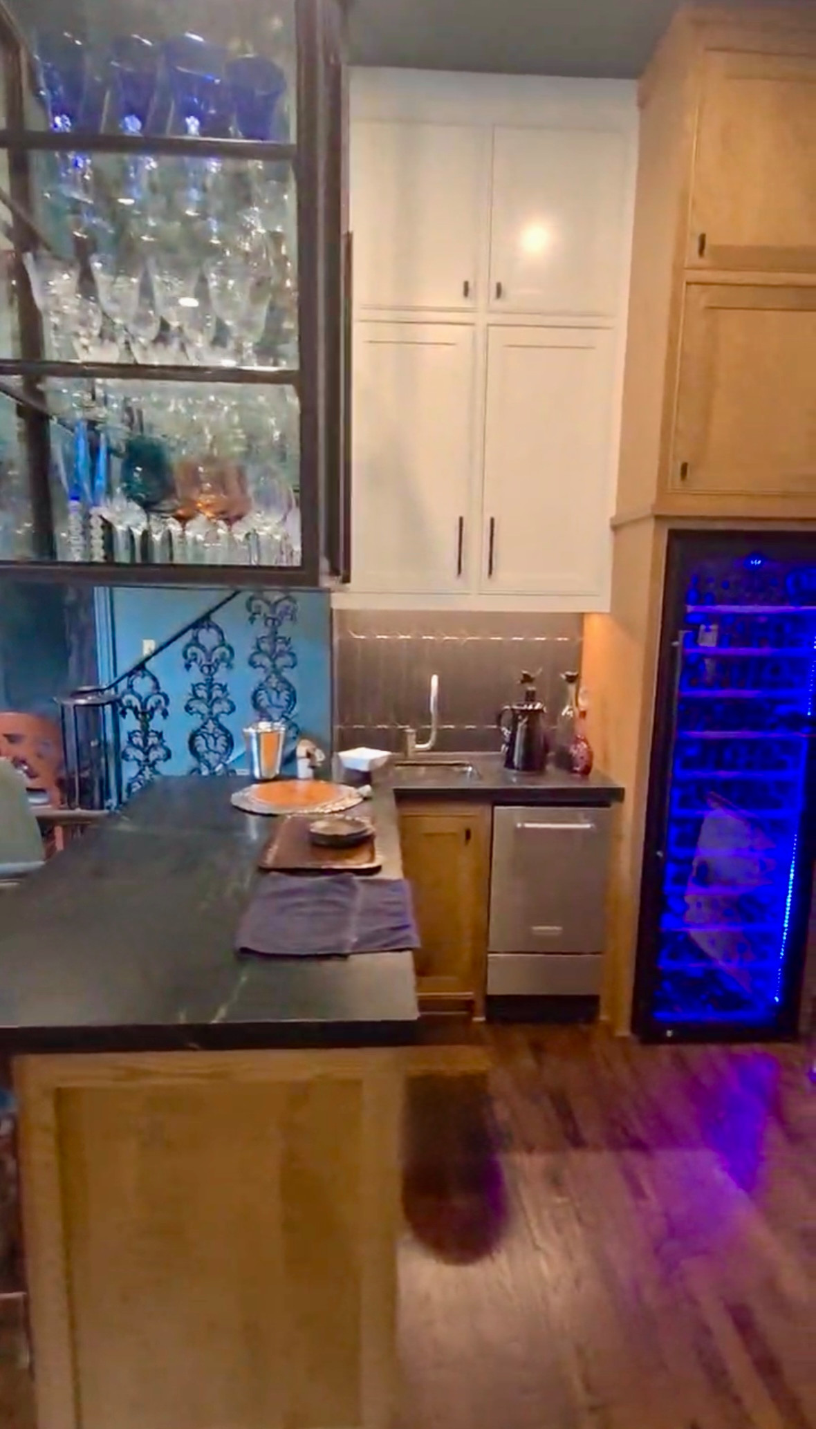 Bar area featuring a full-height wine refrigerator with blue LED lighting, a wet bar sink, and a ceiling-hung metal glass display rack with integrated lighting.