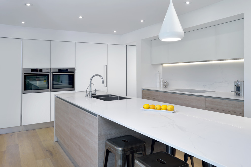 Photo of a contemporary l-shaped kitchen in Toronto with a double-bowl sink, flat-panel cabinets, white cabinets, marble benchtops, white splashback, stone slab splashback, stainless steel appliances, light hardwood floors and with island.