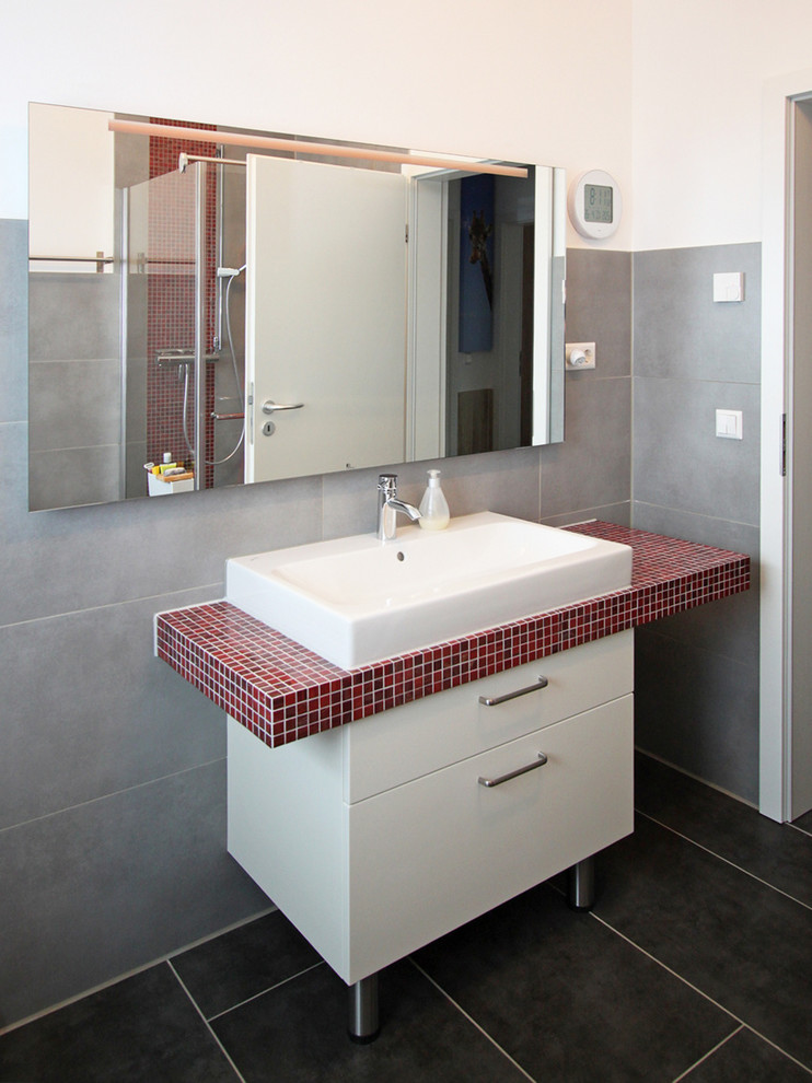 Inspiration for a mid-sized contemporary bathroom in Hamburg with white walls, a vessel sink, tile benchtops, flat-panel cabinets, white cabinets, gray tile and black tile.