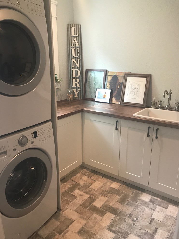 This is an example of a small country l-shaped utility room in Tampa with a drop-in sink, shaker cabinets, white cabinets, wood benchtops, grey walls, brick floors, a stacked washer and dryer and beige floor.