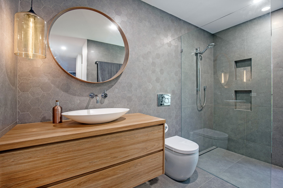 Design ideas for a mid-sized contemporary 3/4 bathroom in Sunshine Coast with medium wood cabinets, gray tile, porcelain tile, grey walls, porcelain floors, a vessel sink, wood benchtops, grey floor, an open shower, flat-panel cabinets, a curbless shower, a one-piece toilet and brown benchtops.