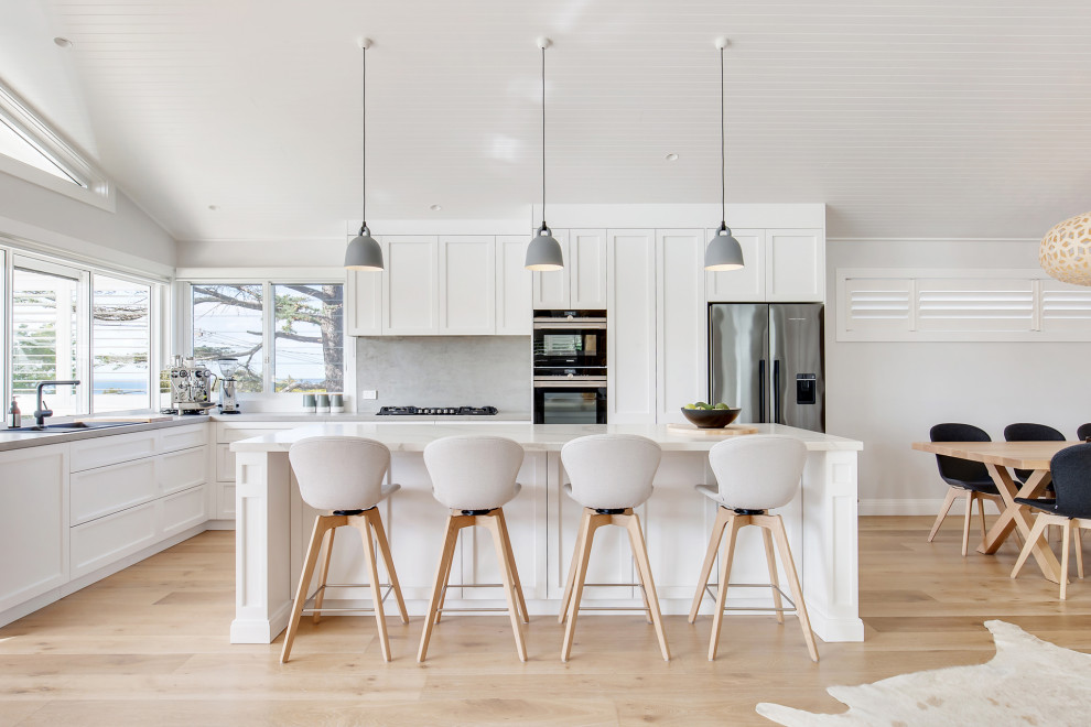 Photo of a large beach style l-shaped open plan kitchen in Sydney with a drop-in sink, shaker cabinets, white cabinets, quartz benchtops, multi-coloured splashback, engineered quartz splashback, stainless steel appliances, light hardwood floors, with island, multi-coloured floor and multi-coloured benchtop.