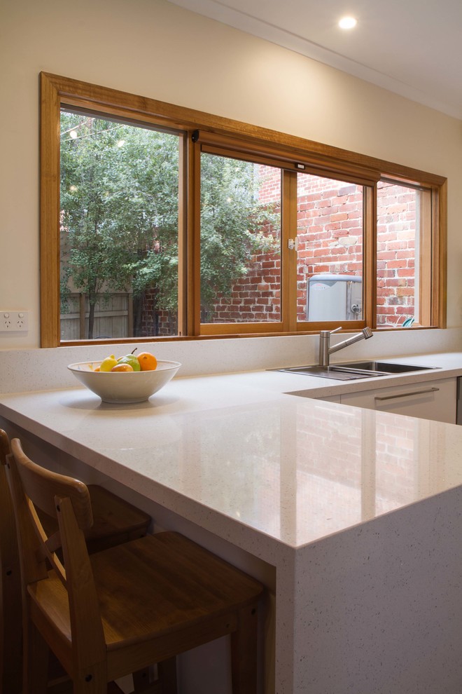Mid-sized contemporary u-shaped separate kitchen in Melbourne with a drop-in sink, flat-panel cabinets, white cabinets, quartz benchtops, metallic splashback, glass sheet splashback, stainless steel appliances, medium hardwood floors and a peninsula.