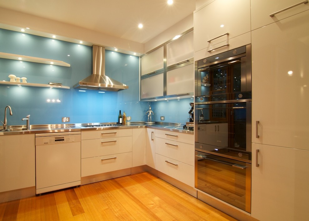This is an example of a mid-sized modern l-shaped separate kitchen in Sydney with an integrated sink, flat-panel cabinets, white cabinets, stainless steel benchtops, blue splashback, glass sheet splashback, stainless steel appliances, light hardwood floors, with island and white benchtop.