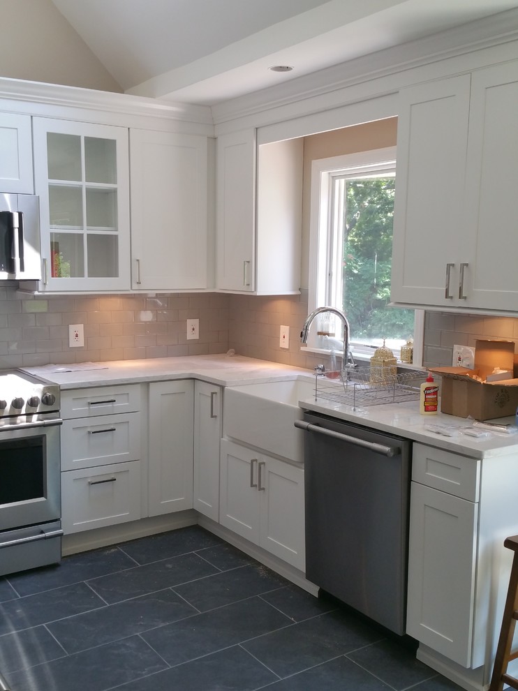 Photo of a mid-sized transitional l-shaped eat-in kitchen in New York with a farmhouse sink, shaker cabinets, white cabinets, quartz benchtops, grey splashback, porcelain splashback, stainless steel appliances, slate floors and grey floor.