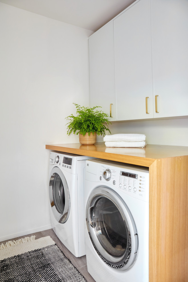 Idéer för små funkis linjära brunt tvättstugor enbart för tvätt, med luckor med infälld panel, vita skåp, träbänkskiva, vitt stänkskydd, vita väggar, klinkergolv i porslin, en tvättmaskin och torktumlare bredvid varandra och grått golv