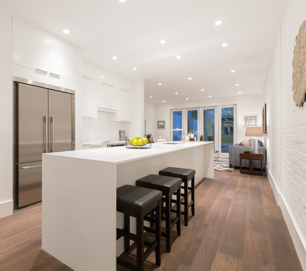 This is an example of a contemporary single-wall eat-in kitchen in DC Metro with flat-panel cabinets, white cabinets, solid surface benchtops, white splashback, stainless steel appliances, dark hardwood floors, brown floor and white benchtop.