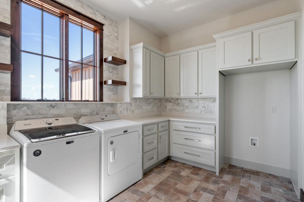 This is an example of an expansive country u-shaped utility room in Dallas with a double-bowl sink, raised-panel cabinets, white cabinets, quartzite benchtops, grey splashback, porcelain splashback, grey walls, brick floors, a side-by-side washer and dryer, red floor and white benchtop.