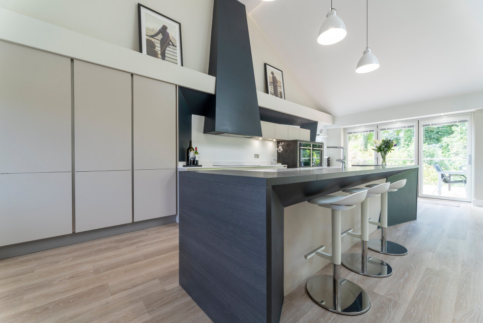 Large contemporary galley open plan kitchen in Edinburgh with a built-in sink, flat-panel cabinets, white cabinets, quartz worktops, white splashback, stone slab splashback, black appliances, light hardwood flooring, an island, beige floors and white worktops.