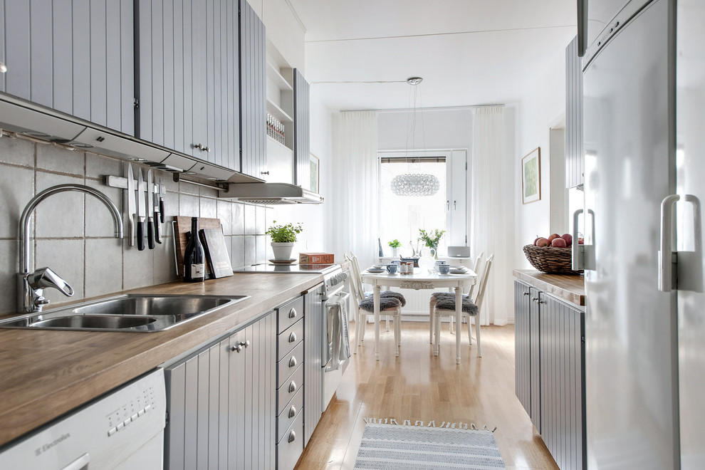 Scandinavian galley eat-in kitchen in Gothenburg with a double-bowl sink, grey cabinets, wood benchtops, white splashback, white appliances, light hardwood floors, no island, beige floor and flat-panel cabinets.