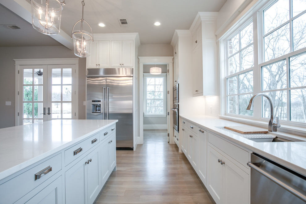 Large traditional u-shaped eat-in kitchen in Dallas with shaker cabinets, white cabinets, granite benchtops, white splashback, glass tile splashback, stainless steel appliances, light hardwood floors, with island and an undermount sink.