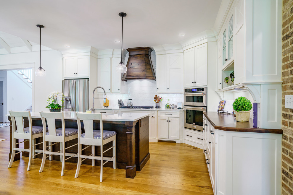 Example of a large transitional u-shaped medium tone wood floor and brown floor eat-in kitchen design with a farmhouse sink, recessed-panel cabinets, white cabinets, quartz countertops, white backsplash, ceramic backsplash, stainless steel appliances, an island and white countertops