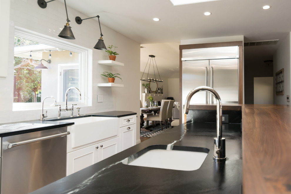 Inspiration for a huge transitional l-shaped medium tone wood floor and brown floor open concept kitchen remodel in San Francisco with a farmhouse sink, recessed-panel cabinets, white cabinets, soapstone countertops, white backsplash, stainless steel appliances, an island and black countertops