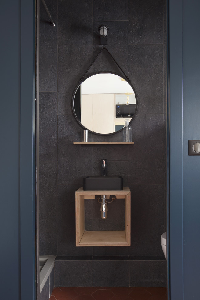Inspiration for a small contemporary powder room in Paris with open cabinets, light wood cabinets, black tile, black walls, a vessel sink, wood benchtops, brown floor, a wall-mount toilet, cement tile and terra-cotta floors.