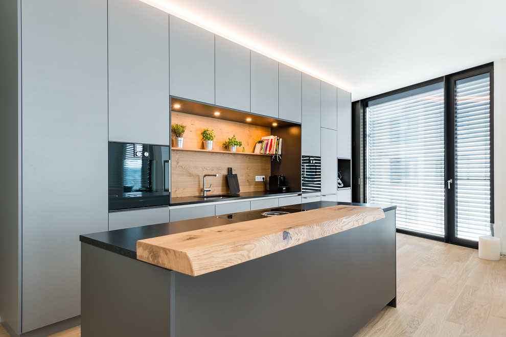 Photo of a small contemporary galley open plan kitchen in Munich with grey cabinets, timber splashback, black appliances, brown floor, black benchtop, an undermount sink, flat-panel cabinets, granite benchtops, brown splashback, medium hardwood floors and with island.