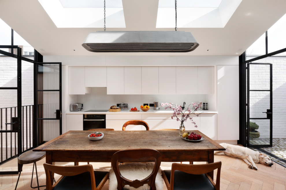 Photo of a contemporary single-wall eat-in kitchen in London with flat-panel cabinets, white cabinets, white splashback, black appliances, light hardwood floors, no island, beige floor and white benchtop.