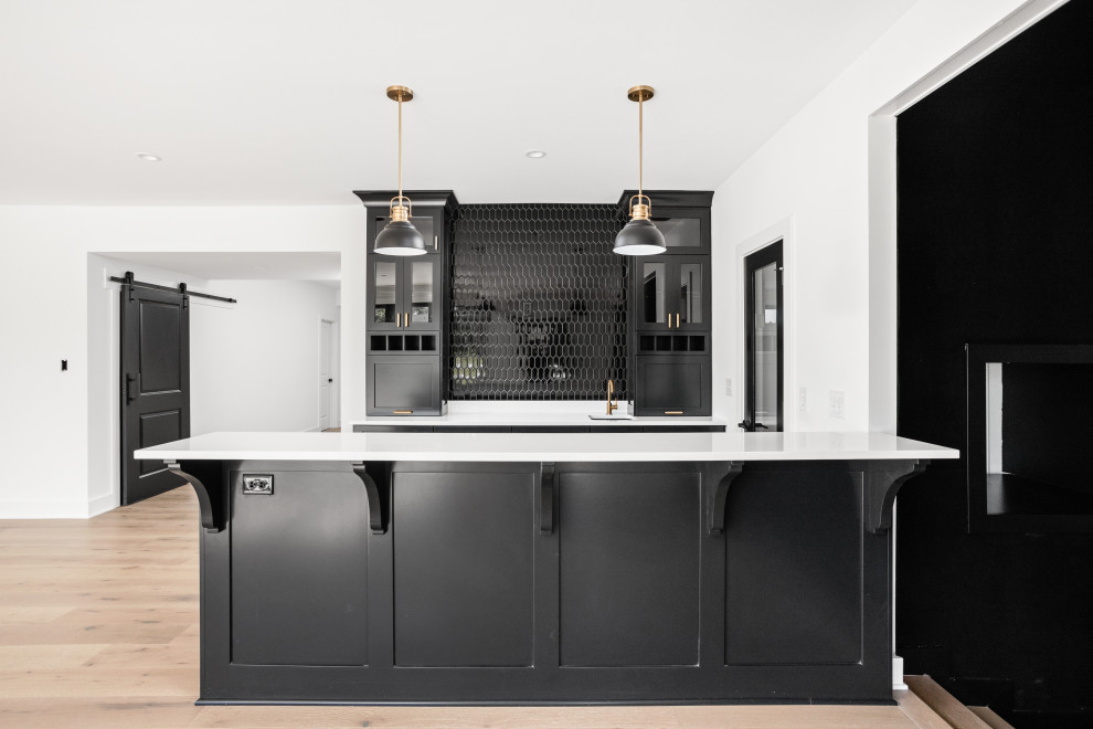 Photo of a mid-sized transitional galley seated home bar in Indianapolis with an undermount sink, shaker cabinets, black cabinets, quartz benchtops, black splashback, ceramic splashback, light hardwood floors, multi-coloured floor and white benchtop.