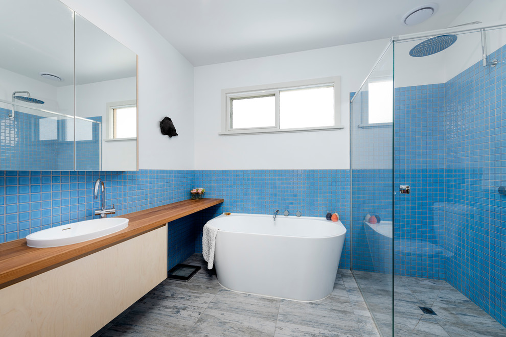 Inspiration for a large beach style master bathroom in Melbourne with flat-panel cabinets, light wood cabinets, wood benchtops, brown benchtops, a freestanding tub, a curbless shower, blue tile, white walls, a vessel sink and grey floor.