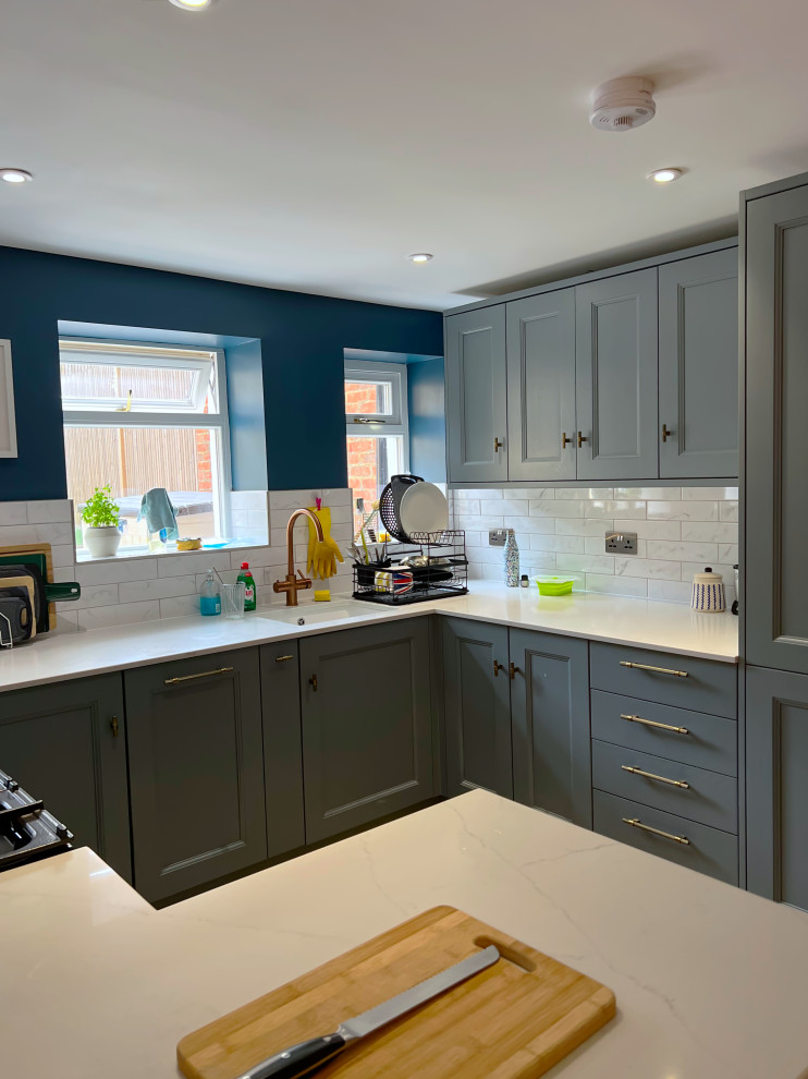 Small contemporary u-shaped eat-in kitchen in London with granite benchtops, with island, light hardwood floors, an integrated sink, recessed-panel cabinets, grey cabinets, white splashback, cement tile splashback, black appliances, brown floor and beige benchtop.