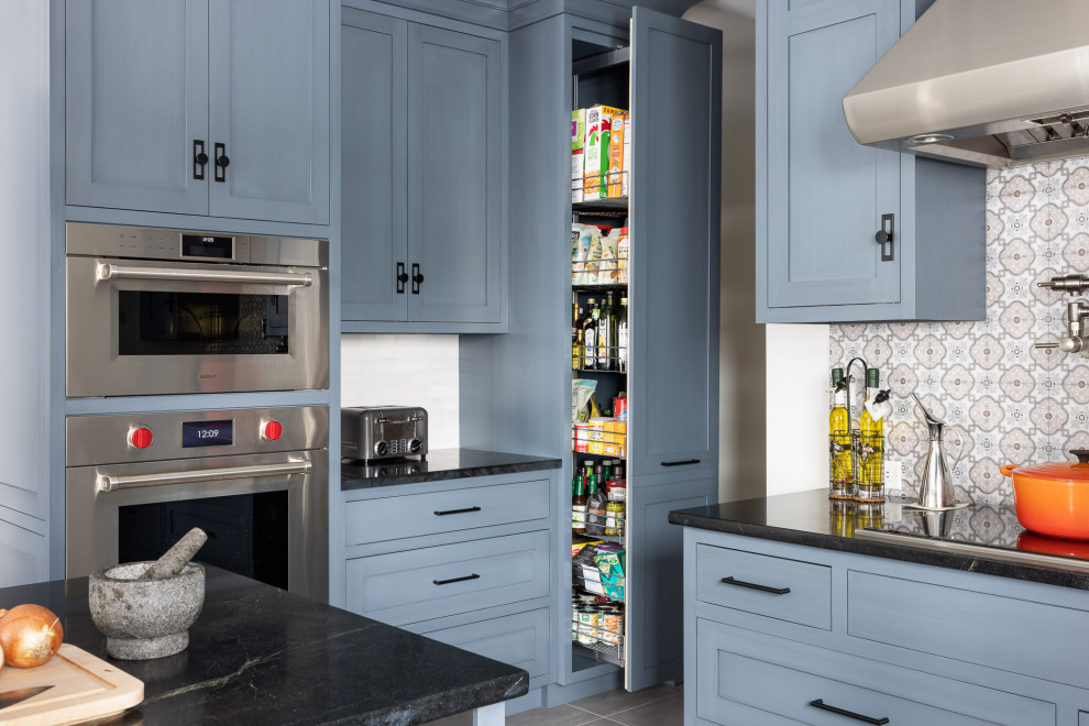 This is an example of a mid-sized eclectic l-shaped kitchen with an undermount sink, recessed-panel cabinets, blue cabinets, soapstone benchtops, white splashback, stainless steel appliances, porcelain floors, with island, grey floor and black benchtop.
